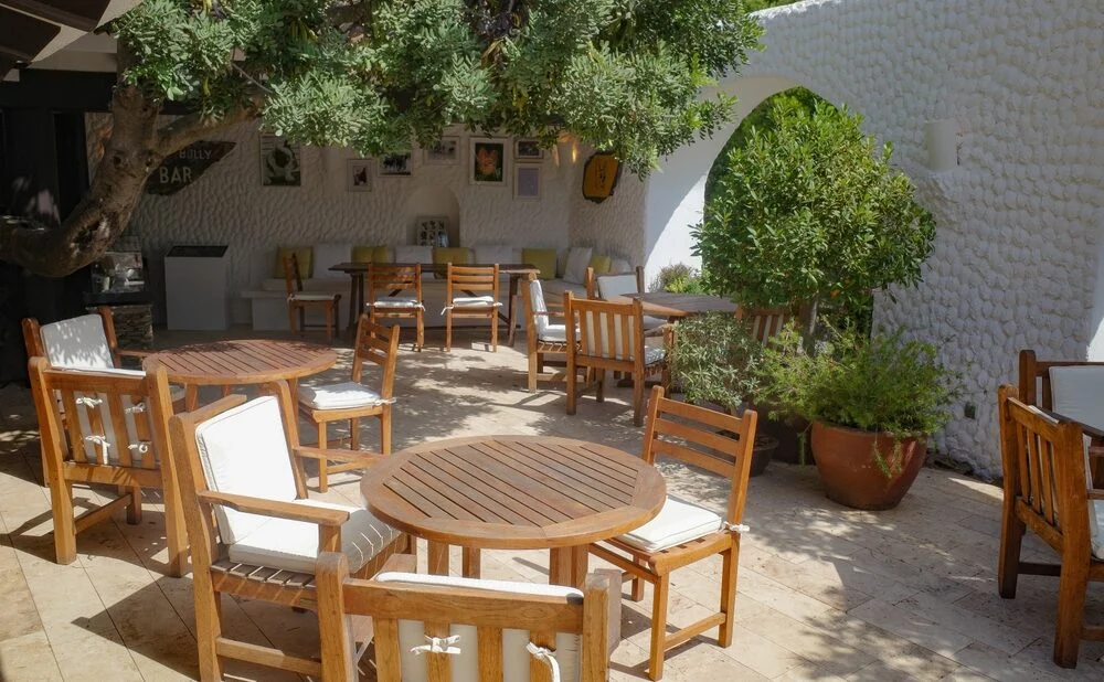 Commercial outdoor patio with wooden dining tables, chairs, and greenery arranged for a restaurant or hospitality space.