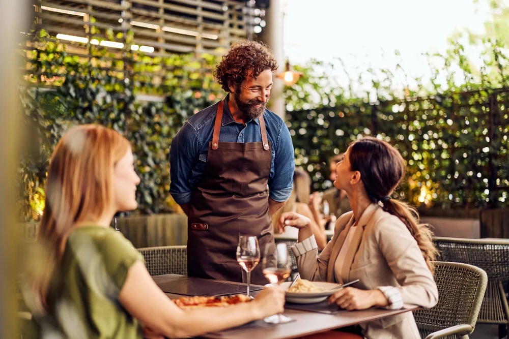 Warm restaurant scene with waiter engaging female, diners outdoor